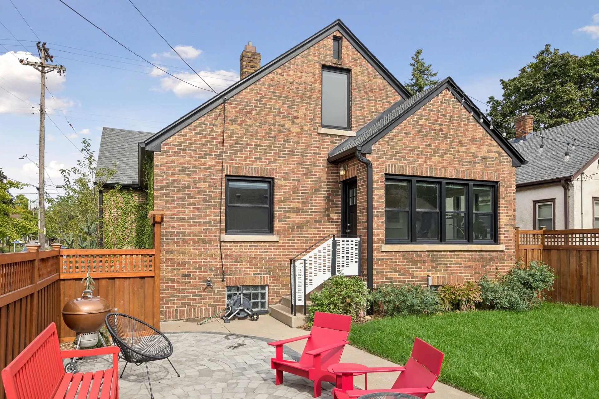 Back of a brick house with black trim, gutters and a downspout, seen across a patio with red chairs and a stained cedar fence
