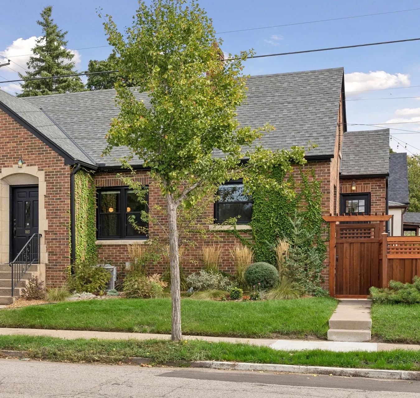 Front of a red-brick house with black trim along the roof edges, a black gutter and downspout, a black front door with iron railings, and a stained cedar gate at the side