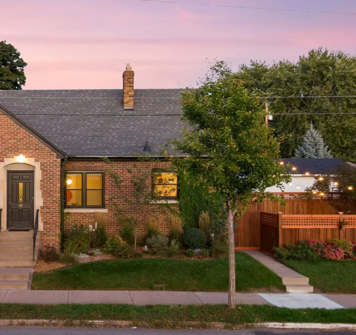Front of a brick house at dusk, the porch light on above a black front door, and string lights over a stained cedar fence at the side